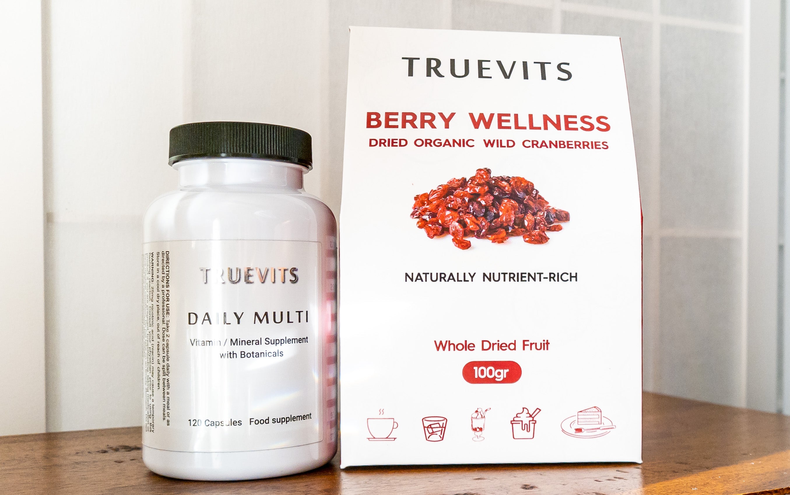 white-bottle-daily-multi-vitamin-mineral-supplement-with-botanicals-standing-on-wooden-console-together-with-berry-wellness-dried-cranberries-100gr-in-white-and-red-paper-box-with-icons-of-cap-on-soucer-ice-cream-piece-of-cake-on-plate